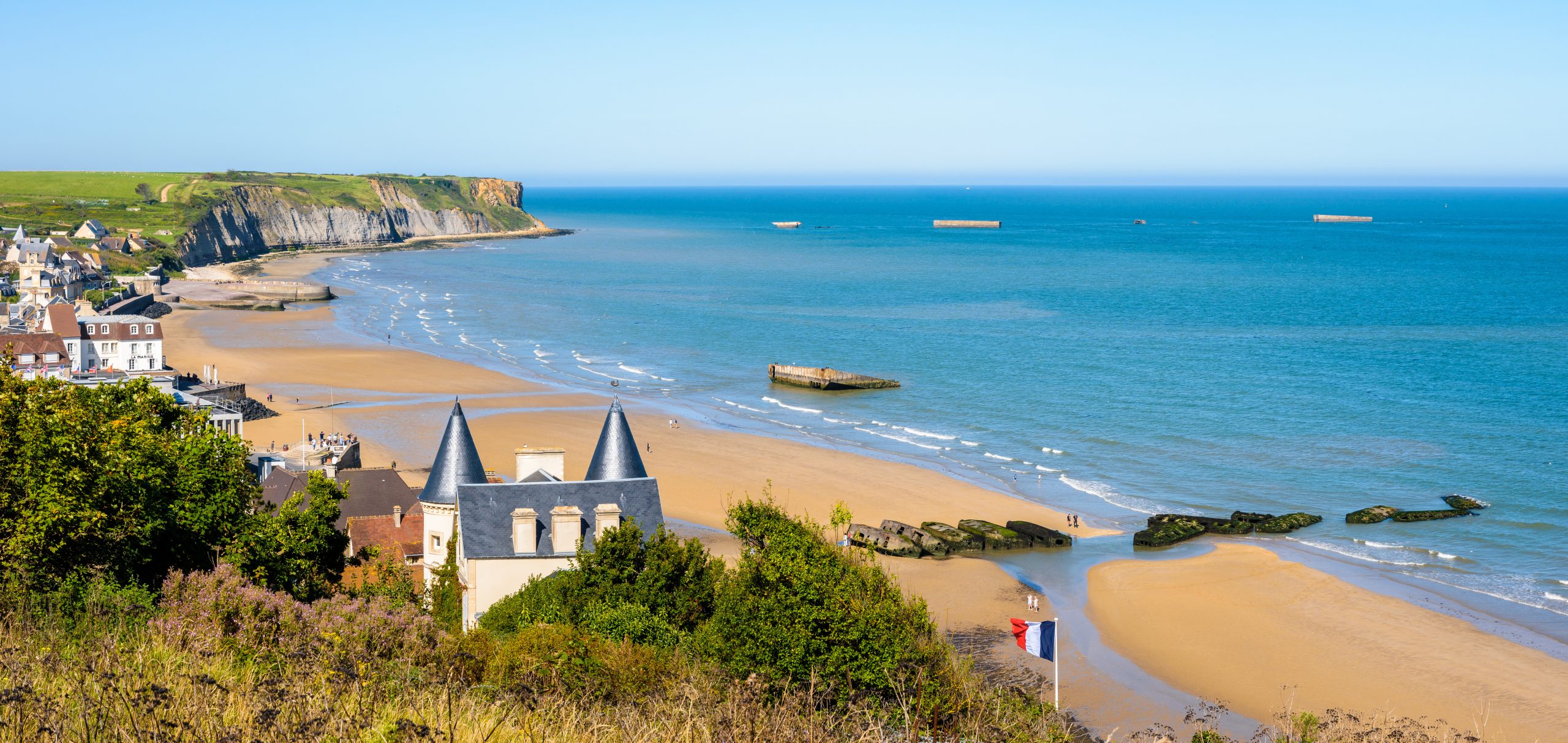 Arromanches beach and Cap Manvieux in Normandy. View of the Normandy coastline with sandy beach, historic buildings, and remnants of World War II structures along the shore.