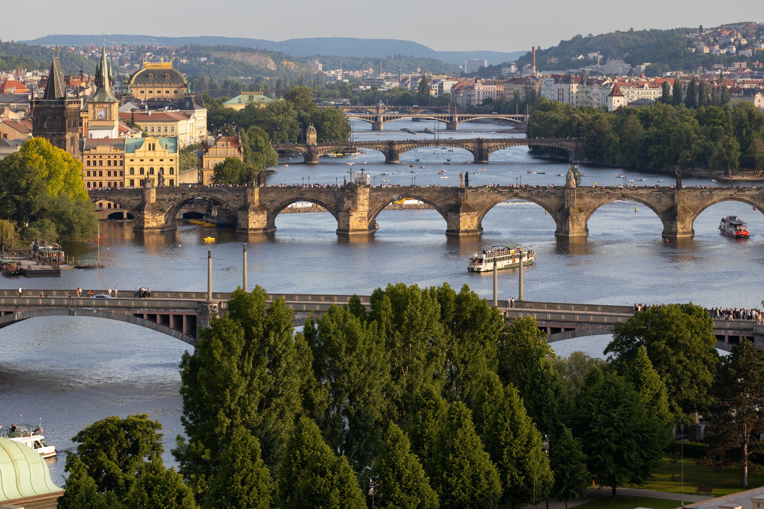 Scenic view of multiple historic bridges spanning the Vltava River in Prague, with lush greenery and city architecture visible along the riverbanks